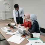 Tim profesional muda berkolaborasi di sekitar meja kantor, menggambarkan delegasi tugas yang efektif